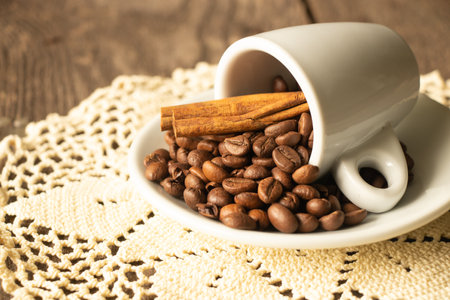 one white cup and saucer with coffee beans and a cinnamon stick stand on a wooden table in the morning in the kitchenの写真素材
