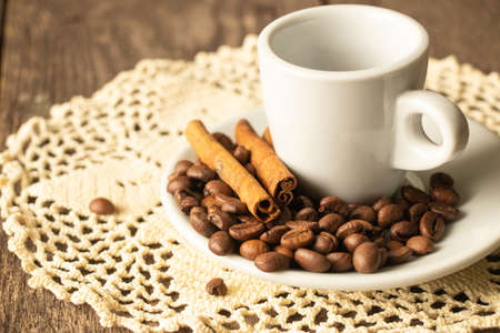one white cup and saucer with coffee beans and a cinnamon stick stand on a wooden table in the morning in the kitchenの写真素材