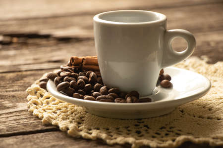 one white cup and saucer with coffee beans and a cinnamon stick stand on a wooden table in the morning in the kitchenの写真素材