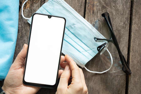 female hand holding a phone with a blank white screen next to glasses and a medical mask on a wooden tableの写真素材