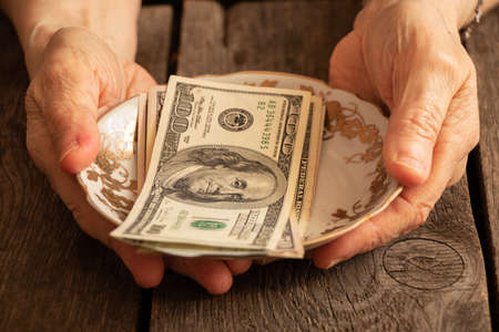 the hands of an elderly woman hold a glass plate with american dollars on a wooden table close-upの写真素材