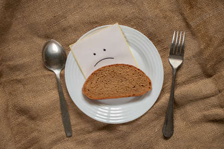 a piece of black bread and a sad smiley face painted next to it on a white plate on the table, forks with a spoon on a brown tablecloth, hunger and nothing to eatの写真素材