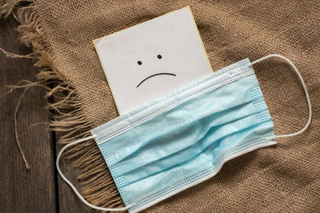 a medical mask and next to a piece of paper with a painted sad smiley face on a wooden table, bad moodの写真素材