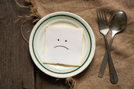 a sad smiley lies on a sheet of paper on a plate next to a spoon and a fork on a brown burlap background, nothing to eat, crisisの写真素材