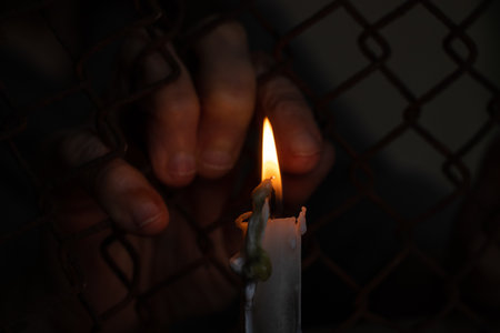 old female hands hang on a fence made of iron mesh in the lock on the background of a burning candleの写真素材