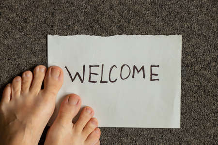 Welcome written on a piece of paper lies on a gray carpet and next to the woman's legs are, welcome...