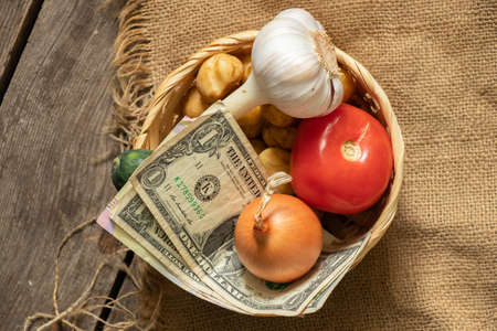 tomato onion garlic potatoes in a wicker basket with dollars on a wooden table, vegetables and moneyの写真素材