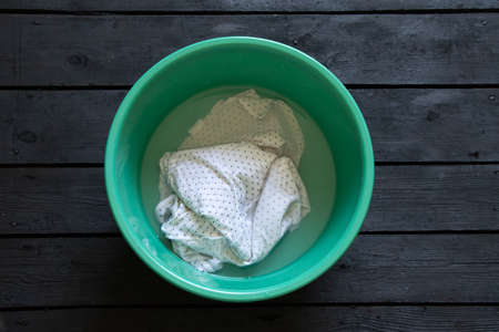 white T-shirt washable in water in a green plastic bowl on a black wooden floor, hand wash in a bowlの写真素材