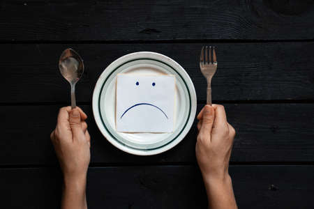 a plate of paper on which a sad smiley face and female hands are drawn, hunger and nothing to eat, a bad mood at lunchの写真素材
