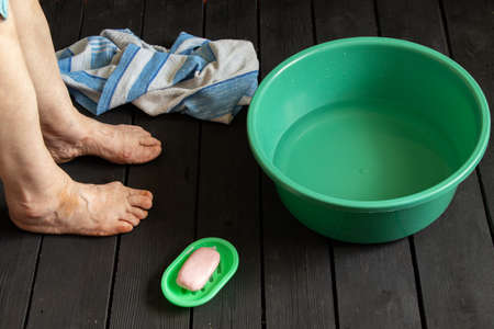 the old grandmother's feet are washed in a plastic bowl with water and soap on the black floor, towels were brought, the old grandmother washes her feet in a bowl on the floor, feet were washedの写真素材