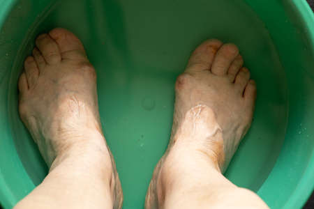 the old grandmother's feet are washed in a plastic bowl with water and soap on the black floor, towels were brought, the old grandmother washes her feet in a bowl on the floor, feet were washedの写真素材
