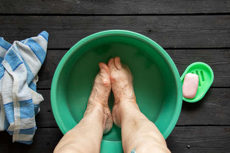 the old grandmother's feet are washed in a plastic bowl with water and soap on the black floor, towels were brought, the old grandmother washes her feet in a bowl on the floor, feet were washedの写真素材