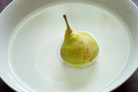 a pear in water in a white mask stands on a table by the window, wash a pear in a plateの写真素材