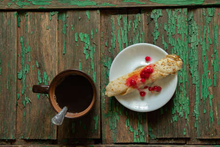 rolled pancake with viburnum and sugar on a teatka next to a cup of coffee on an old wooden table, morning coffee with a dissert on the tableの写真素材