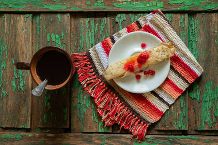 rolled pancake with viburnum and sugar on a teatka next to a cup of coffee on an old wooden table, morning coffee with a dissert on the tableの写真素材