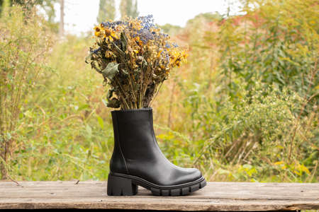 women's high black boots with thick soles stand on a wooden table with a bouquet of wild flowers in the inside of the shoe, women's winter shoes, footwear and fashionの写真素材