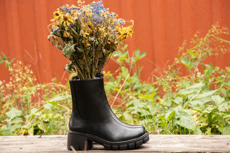 women's high black boots with thick soles stand on a wooden table with a bouquet of wild flowers in the inside of the shoe, women's winter shoes, footwear and fashionの写真素材