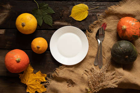 white plate stands on a wooden table next to pumpkins and autumn leaves, festive autumn table setting, dishes on the tableの写真素材