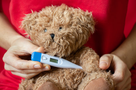girl holding a brown teddy bear with a thermometer in her hand at home in the bedroom on the bed,...