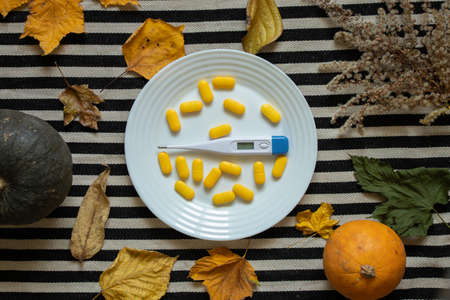 an electronic thermometer lies next to yellow pills on a plate on the table in the kitchen with leaves and pumpkin nearby, medicine and pharmacy, health and treatment, pills and antibiotics seasonal flu diseaseの写真素材