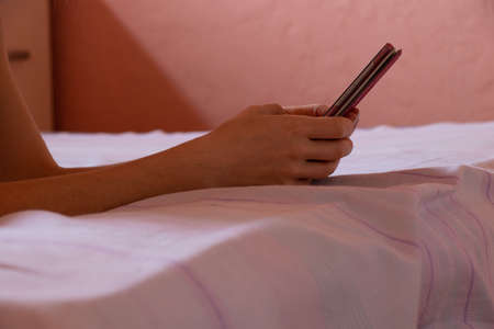 a girl holds a phone in her hands on the bed at night in the bedroom, communication on the phone while sleeping at night on the bed, a phone in her handsの写真素材