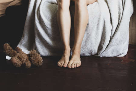 the girl sits on the sofa with her feet on the floor, feet on the wooden floor of the house in the children's room next to a teddy bear, female legs are lying on the couch on the floorの写真素材