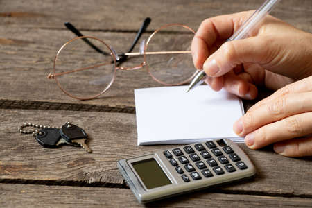 a table and a woman's hand writes with a pen on a sheet of paper next to it lies a calculator, keys and glasses, a workplace, a deskの写真素材