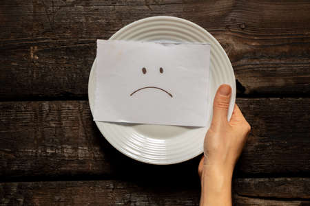 a sad emoticon is drawn on a card that lies on a white plate in the hands of a girl on the table, a bad mood and a crisis, poverty and sadnessの写真素材