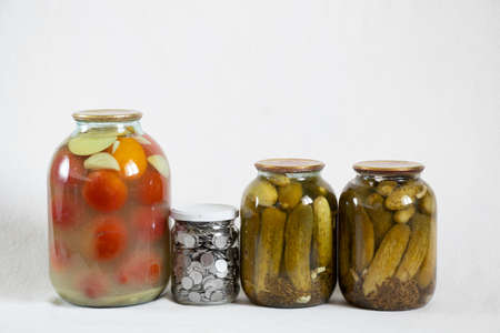 canned tomatoes and cucumbers in jars and next to a bank with Ukrainian kopecks on a white background, save money in a bank, finance and foodの写真素材