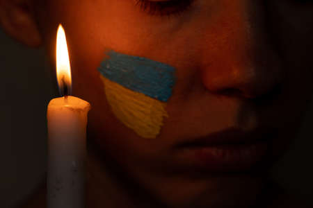 The girl's face with the national flag of Ukraine yellow-blue on the face of the background of the candle flame, patriot, peace in Ukraine, prayer for the country 2022の写真素材