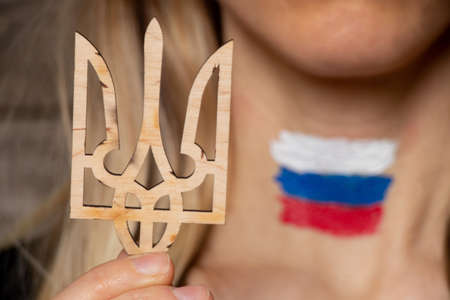 A girl holds a wooden coat of arms of Ukraine against the background of the coat of arms of Russia painted on her neck, Russia's war with peaceful Ukraine, stop the war, peace in Ukraine 2022の写真素材