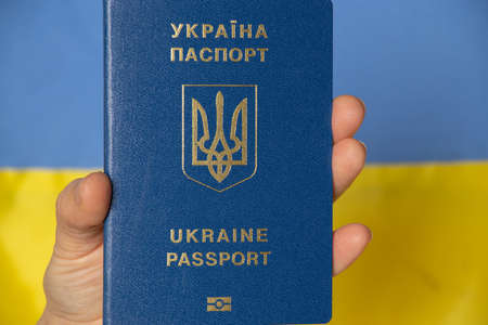 A hand holds a Ukrainian foreign passport against the background of the yellow-blue flag of Ukraine, our peaceful countryの写真素材