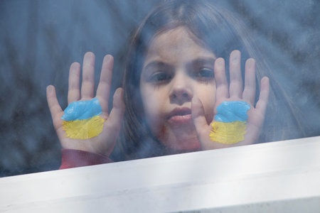 A little girl in the window of her house with a painted yellow-blue flag of Ukraine, peace in Ukraine, stop the war, protest and patriot, home Ukraineの写真素材