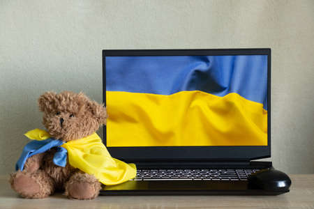 The national flag of Ukraine as a background on a laptop and a teddy bear sits at home next to the flag of Ukraine on the table. Stop the war, patriotism 2022の写真素材