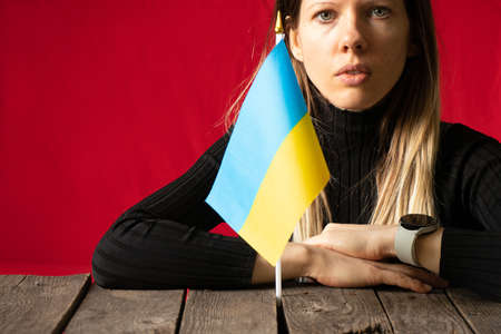 A girl sits at a table at home and holds the national flag of Ukraine against the backdrop of a red wall, stop the war in Ukraine, I am Ukrainian 2022の写真素材