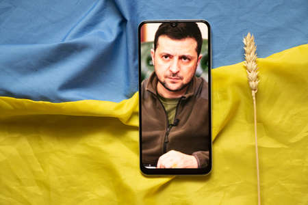 Ukraine Dnipro 01.04.2022 - President of Ukraine Volodymyr Zelensky on the screen of the phone and next to the flag of Ukraine, online news from the president on a smartphone at homeのeditorial素材