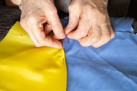 Female old Ukrainian woman hands sew the national flag of Ukraine yellow-blue at the table at home in Ukraine, patriot, stop the war in Ukraine 2022の写真素材