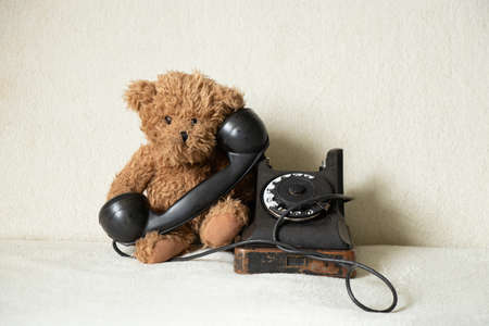 A childish brown teddy bear sits near an old telephone at home and is sad, a retro telephone on a white backgroundの写真素材