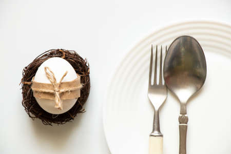 A spoon and a fork lie in a plate, and next to it on the table is a chicken egg, decorated with a rope for the Easter holiday, table decoration for the Easter holiday, cutlery in the kitchenの写真素材