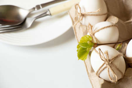 Knife and fork in a plate on a white table and next to a tray with chicken eggs decorated with a...