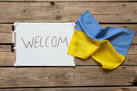 A white sign with the text welcome and next to it lies the flag of Ukraine on a wooden table, EU hospitality for Ukrainiansの写真素材