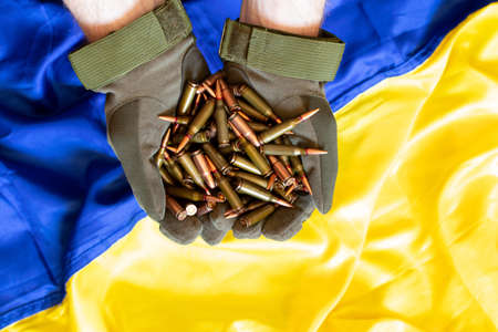 A Ukrainian soldier in gloves holds bullets from a machine gun against the background of the flag of...