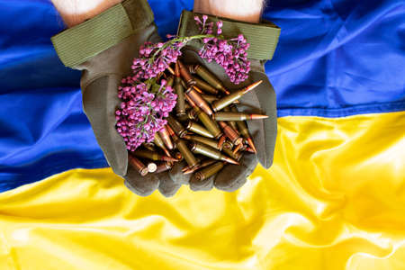 A Ukrainian soldier in gloves holds bullets from a machine gun against the background of the flag of...