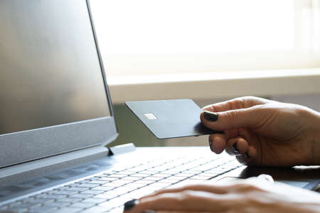 A girl holds a credit bank card in her hands near a black laptop in an office near a window, online payment, internet card paymentの写真素材