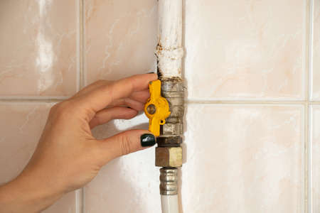 The hand closes the faucet on the gas pipe against the background of the tiles of the house in the apartment, the faucet on the gas pipe in the kitchenの写真素材