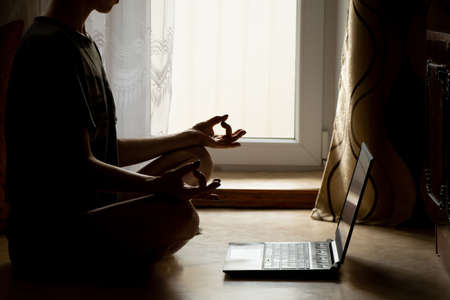 A girl is doing yoga near a laptop on the floor of a house near a window in the dark, an online video lesson on meditation, yoga and miditatsiの写真素材