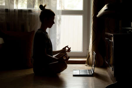 A girl is doing yoga near a laptop on the floor of a house near a window in the dark, an online video lesson on meditation, yoga and miditatsiの写真素材