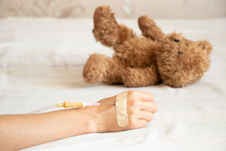 Children's hand with a dropper on the bed and next to it lies a children's teddy bear in the hospital in the ward, children's medicine, a sick childの写真素材