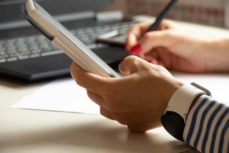 A girl in shirts sits at a table with a phone in her hands, near a laptop, near a window in the...