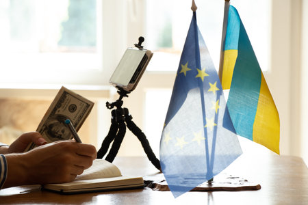 A girl with dollars in her hands is talking on a video chat on the phone at the table, finance and success, the flag of Ukraine and the European Union are on the table, international negotiationsの写真素材